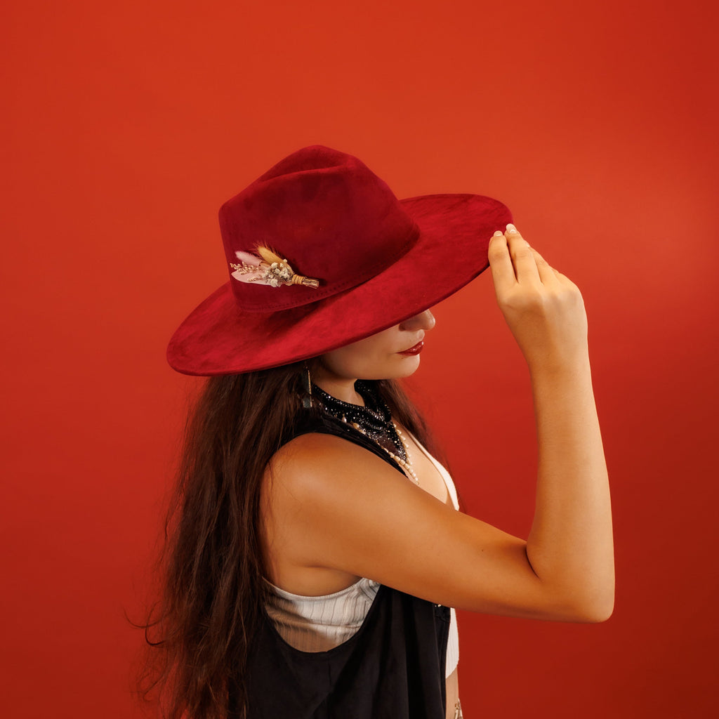 A woman in profile wearing a wide-brimmed red hat with a feather accent, posed against a solid red background.