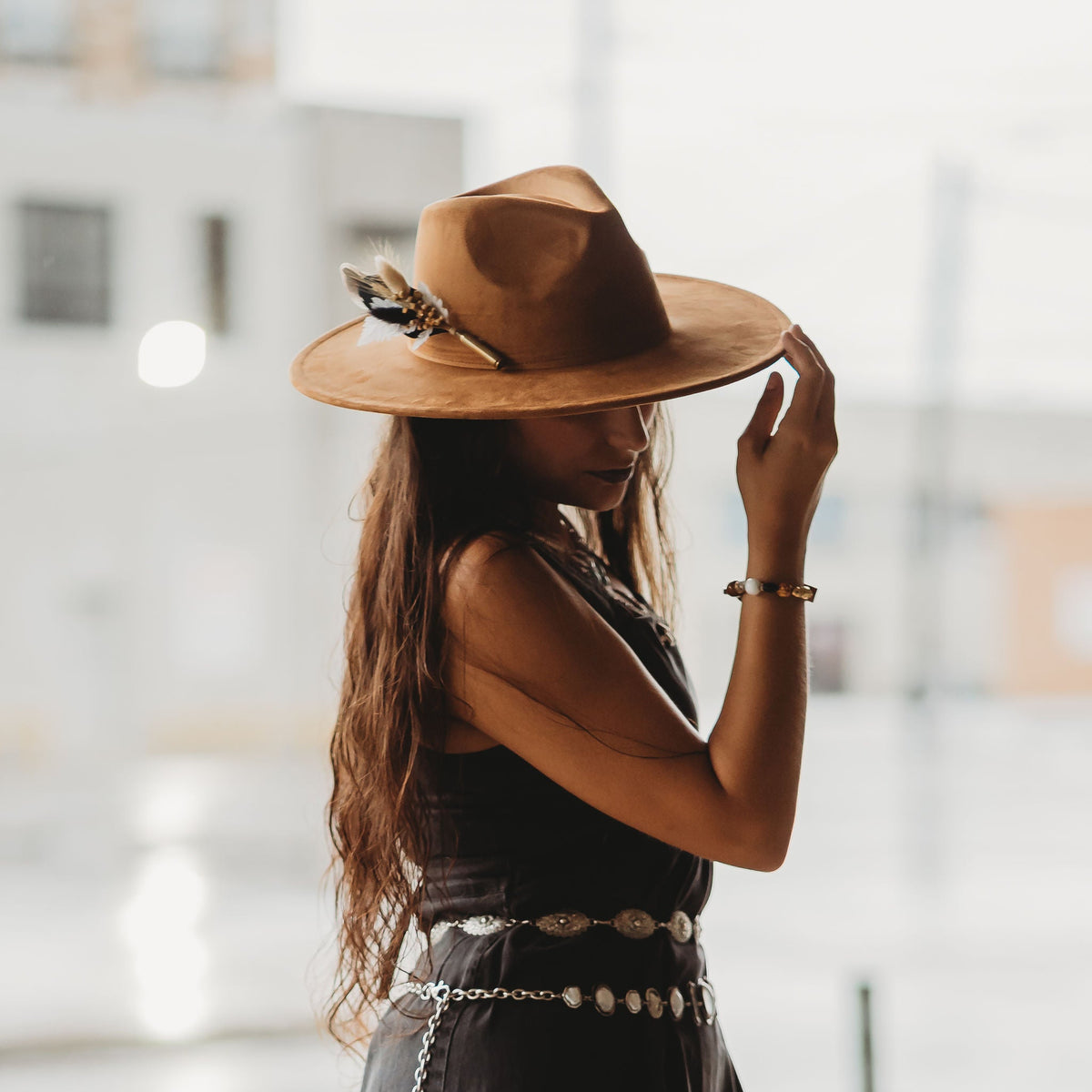 A woman in a black sleeveless dress with a silver chain belt and a brown wide-brim hat with a feather BrimBud accent, standing in a bright, softly blurred outdoor setting.