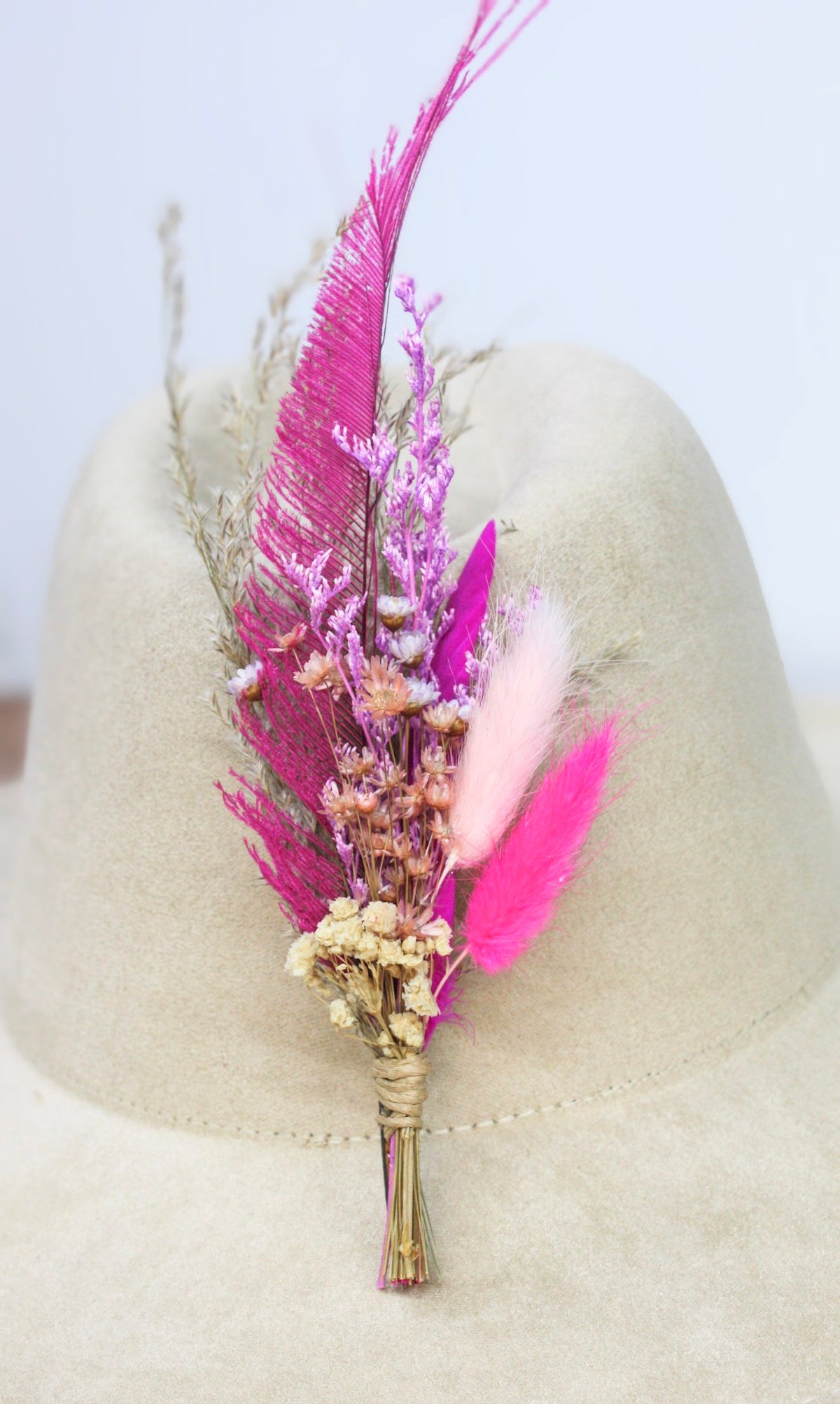 A dried flower and feather hat pin featuring vibrant magenta and pink feathers, light pink pampas grass, and small beige dried flowers. 