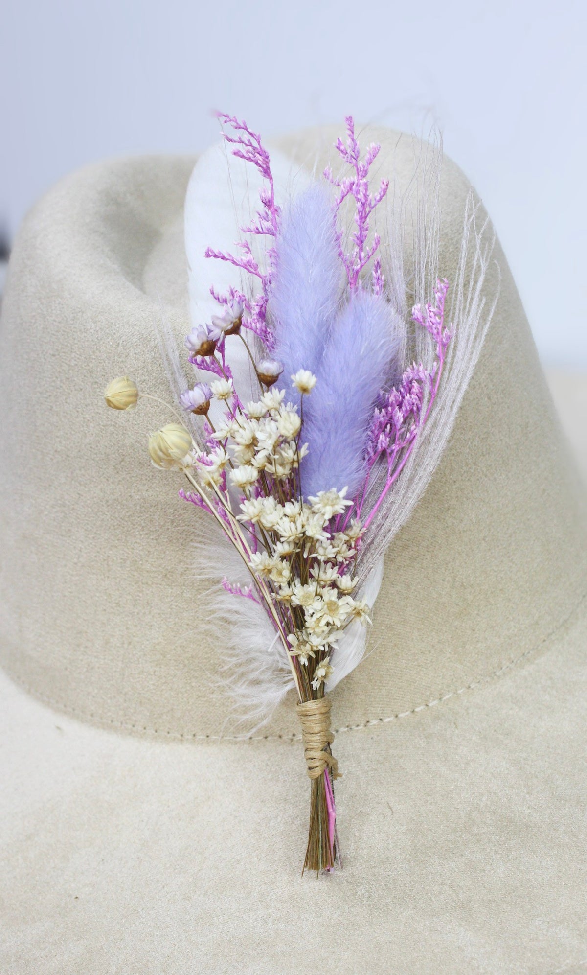 A dried flower and feather hat pin featuring lavender feathers, light purple pampas grass, and small beige dried flowers. 