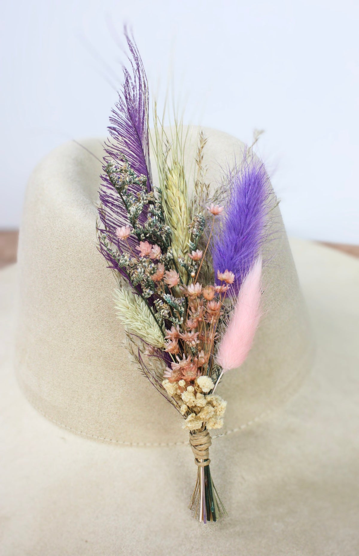 A dried floral hat pin featuring vibrant purple and pink feathers, small pink and purple flowers, and grasses, wrapped in natural twine and displayed on a light beige felt hat.