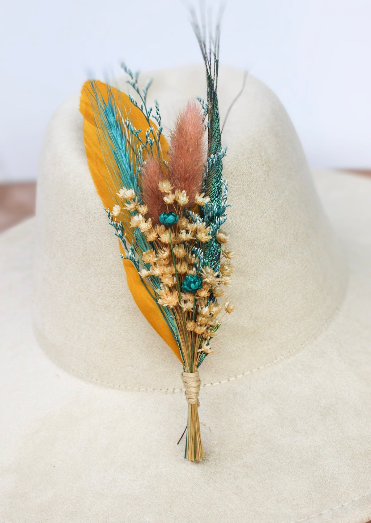 A bohemian-style feather and dried flower hairpiece accessory with a large mustard yellow feather, teal feather, pink pampas grass, and small cream flowers, displayed on a wide-brimmed cream felt hat.