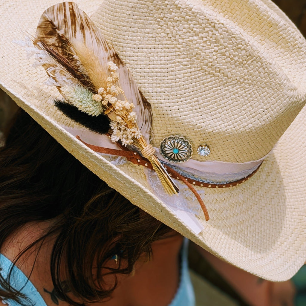A close-up of a person wearing a light beige woven cowboy hat decorated with a brown feather, dried floral accents, and a patterned band, paired with a light blue top.