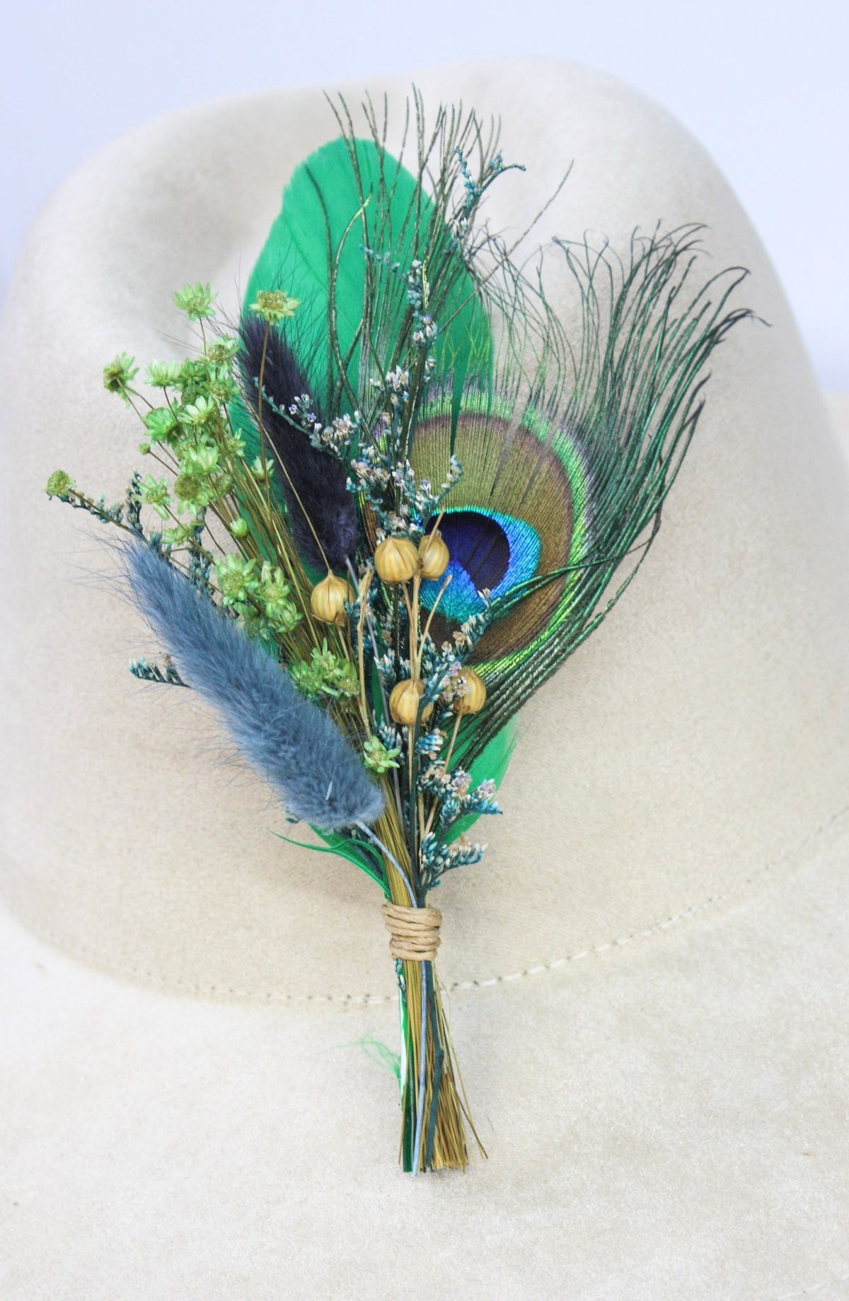 A dried floral hat pin featuring a vibrant green peacock feather, blue pampas grass, small white flowers, and gold berries, arranged on a light beige felt surface.