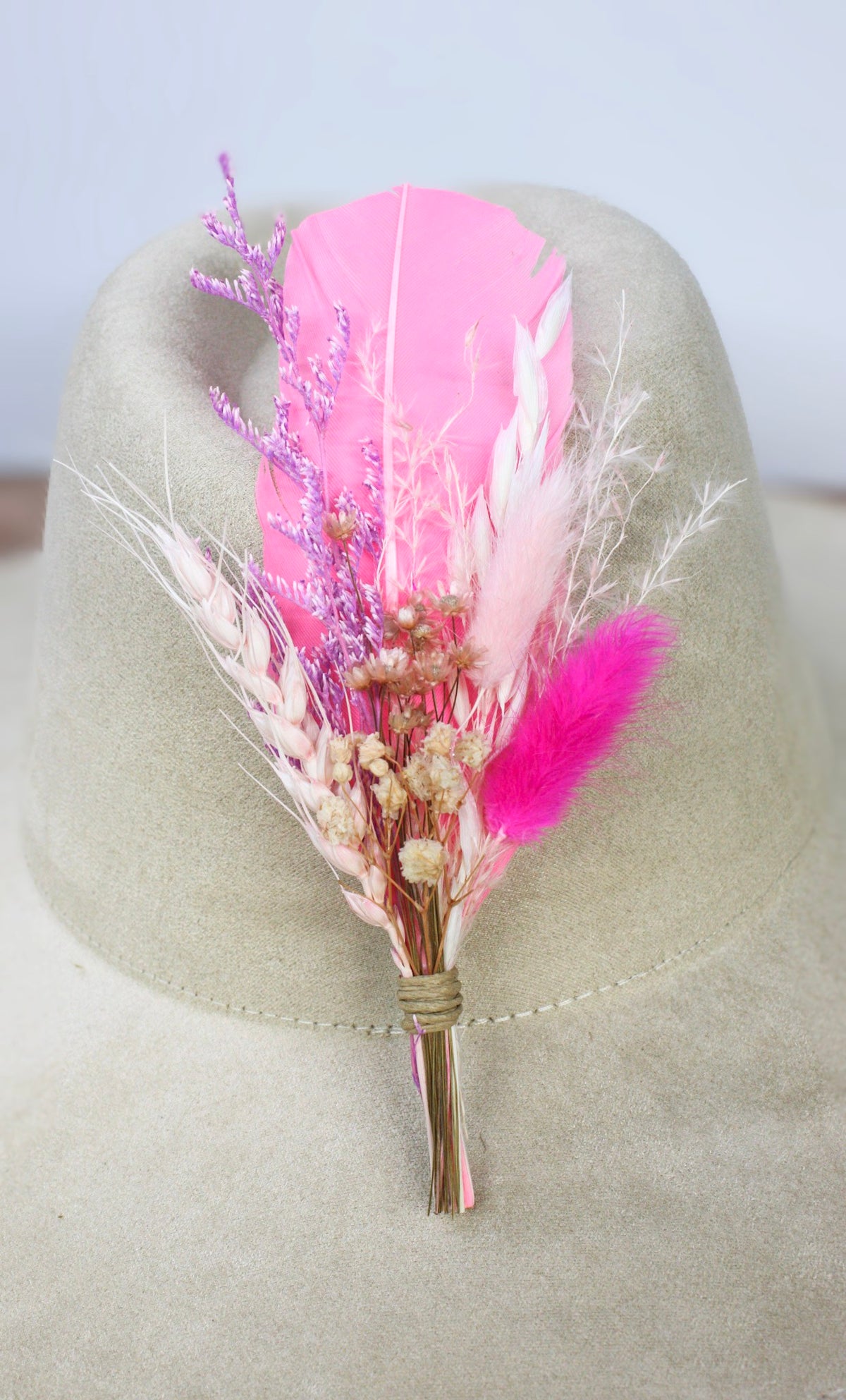 A dried floral hat pin featuring vibrant pink feathers, small pink, white and cream flowers, and light pink grasses, wrapped in natural twine and displayed on a light beige felt hat.