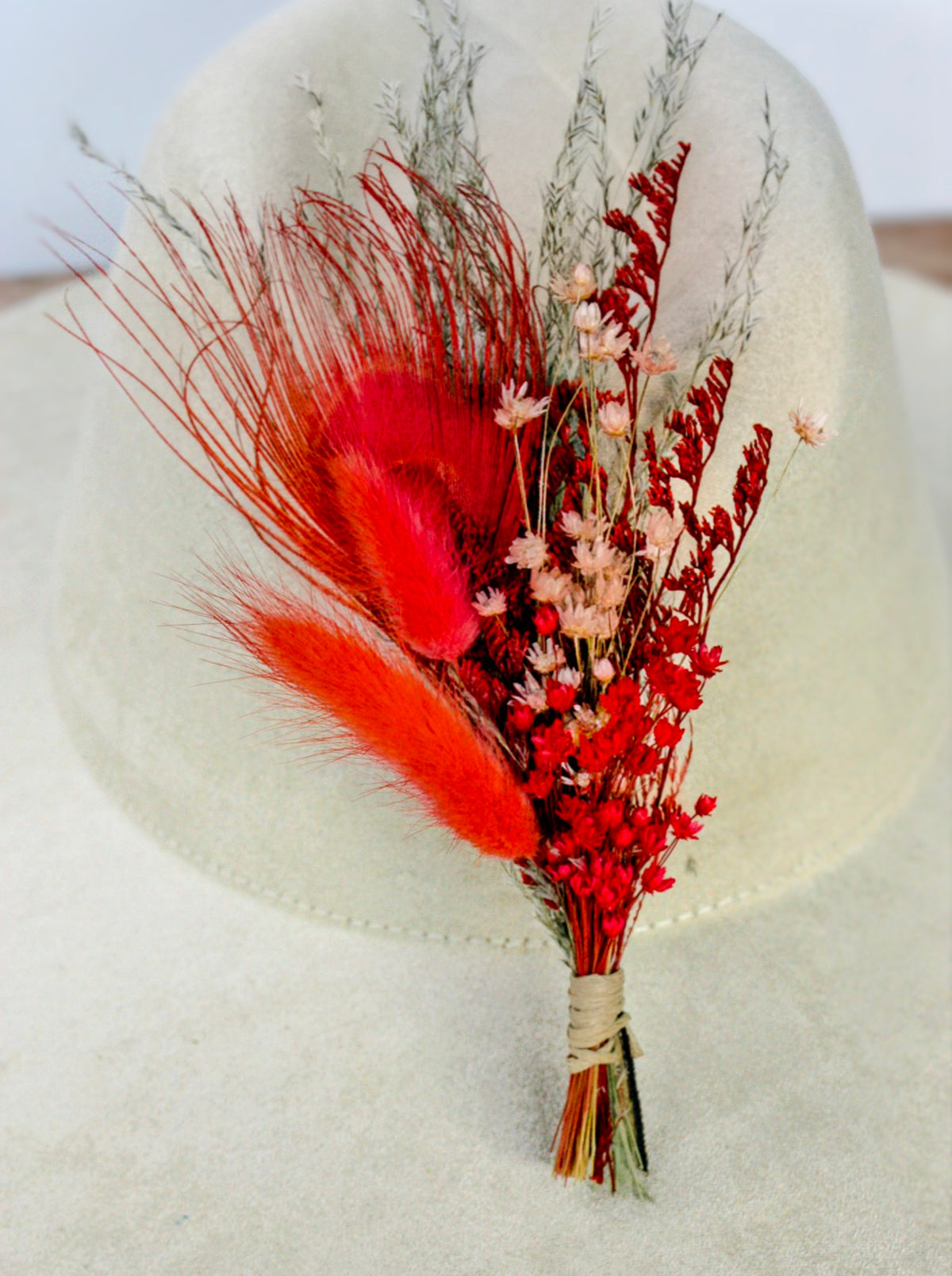 A dried floral hat pin featuring vibrant red feathers, small red and pink flowers, and silver-gray grasses, wrapped in natural twine and displayed on a light beige felt hat.