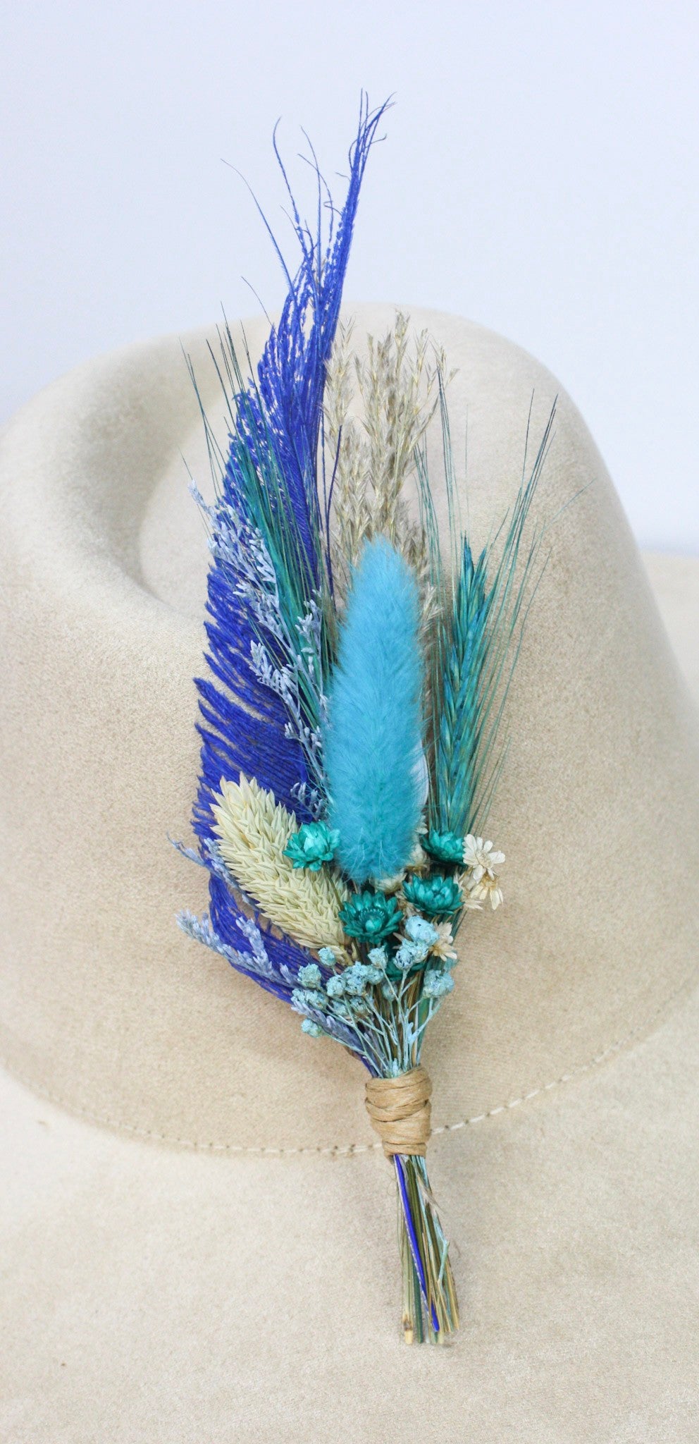 A decorative dried floral and feather hat pin with blue, teal, and beige botanicals, displayed on a beige felt hat.