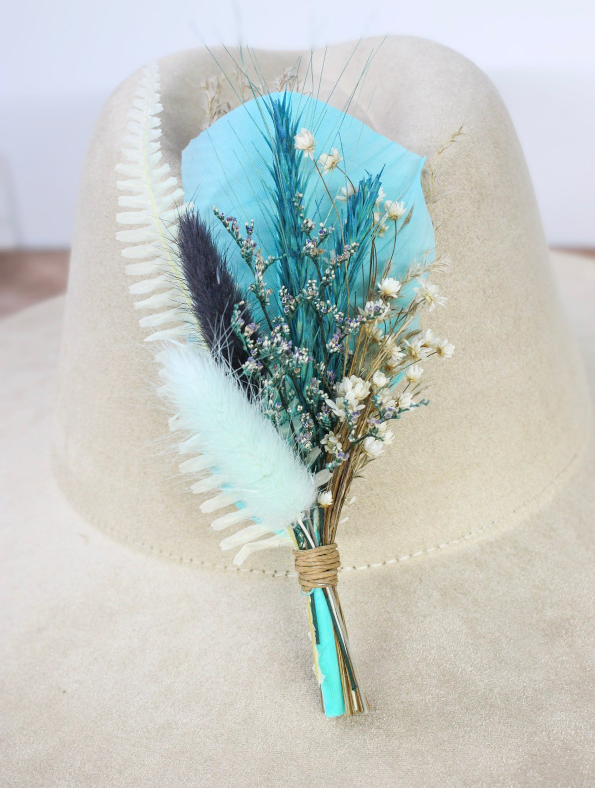 A dried flower and feather hat pin featuring vibrant blue and white feathers, light blue pampas grass, and small dried flowers. 