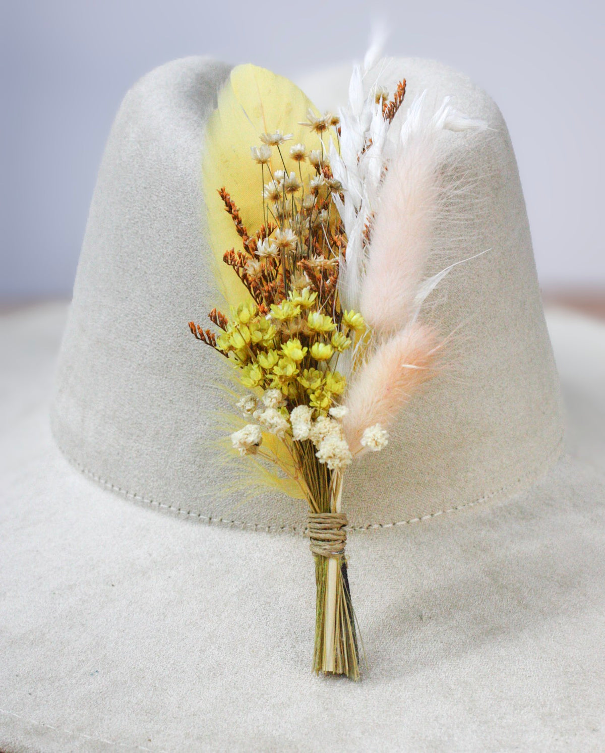 A dried floral hat pin featuring vibrant yellow feathers, small yellow, white and brown flowers, and white grasses, wrapped in natural twine and displayed on a light beige felt hat.