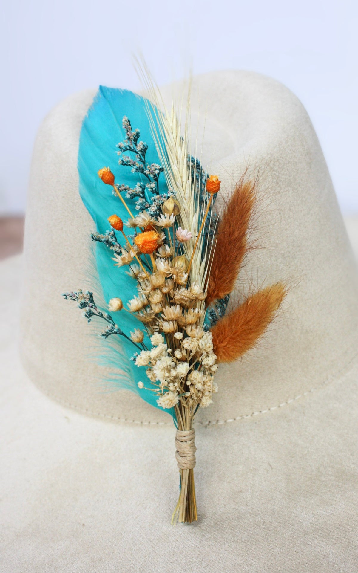 A dried floral hat pin featuring vibrant teal feathers and small orange and cream flowers, wrapped in natural twine and displayed on a light beige felt hat.