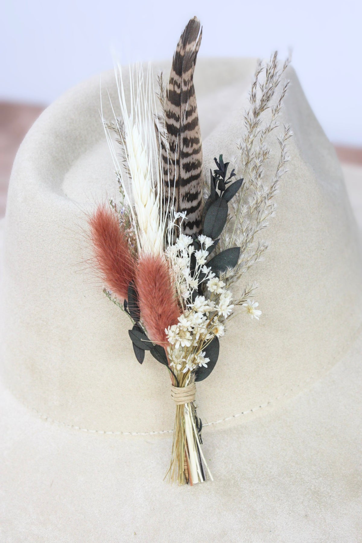A dried floral and feather decorative hat pin displayed on a cream-colored felt hat. The arrangement includes a tall brown-and-black patterned feather, two soft pinkish-brown feathers, white baby's breath flowers, beige dried grasses, and dark green leaves tied together with natural twine.