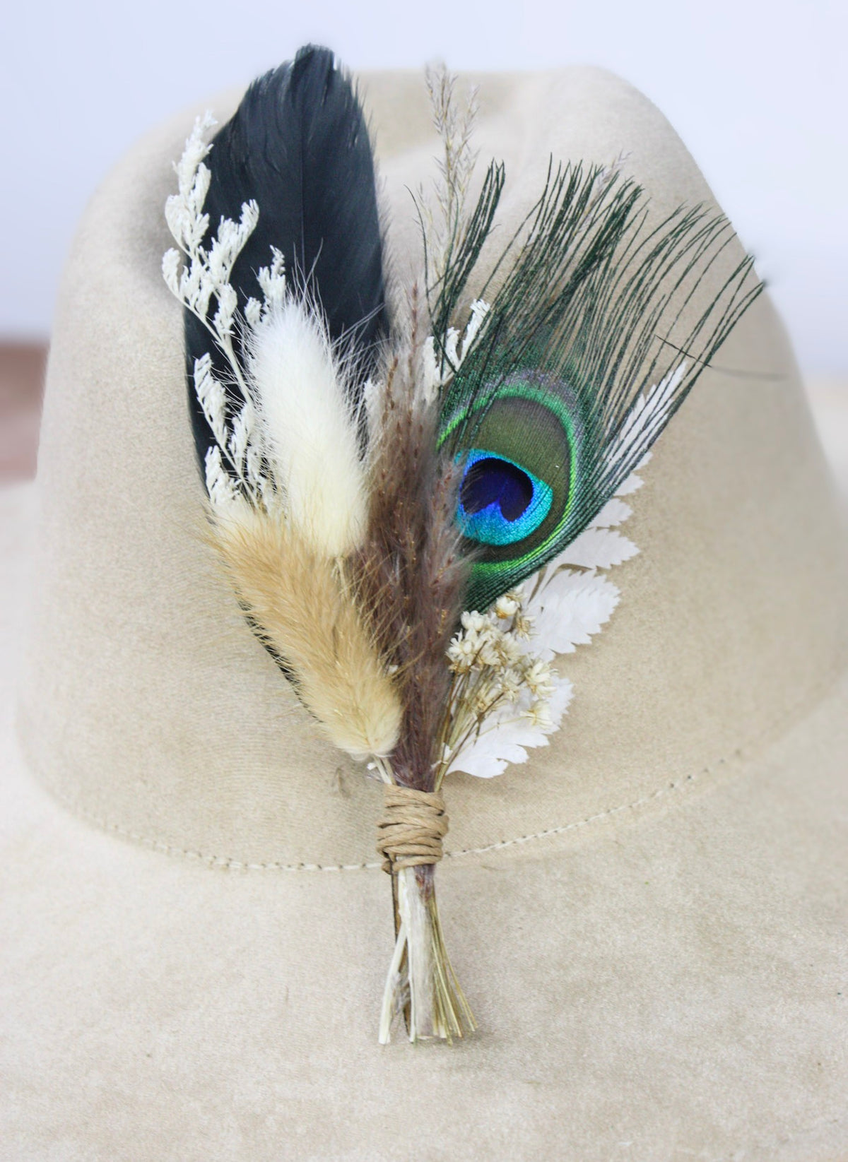 A hat pin displayed on a cream felt hat, featuring a black feather, white dried flowers, a tan feather, and a green peacock feather with blue eye detail.