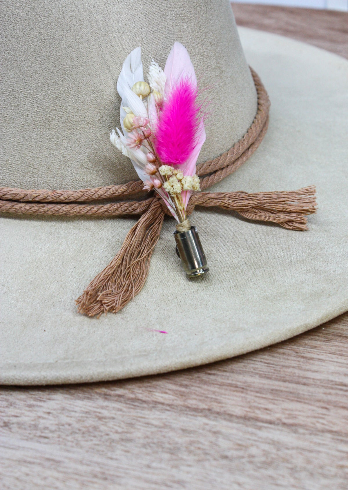A close-up of a beige wide-brim hat with a brown rope band and tassel, decorated with a feathered pin featuring white, pink, and light brown feathers on a wooden surface.