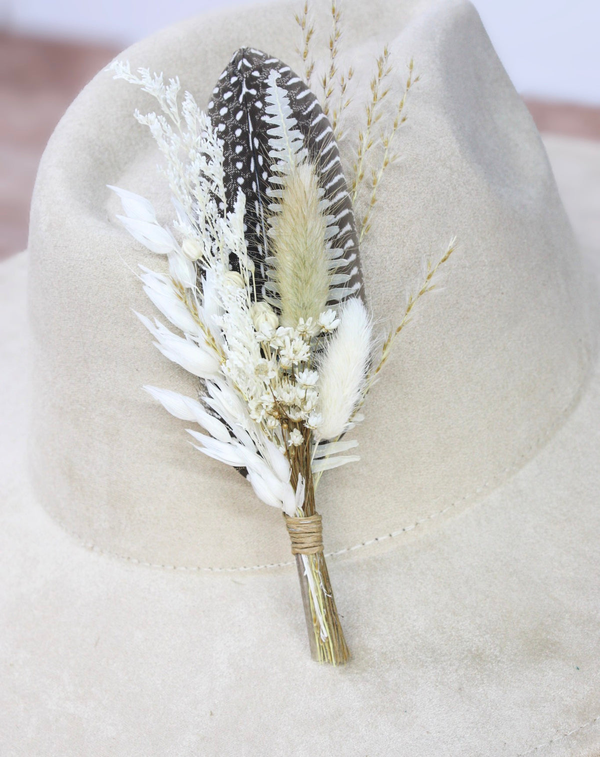 A dried floral hat pin featuring a black and white patterned feather, a tan feather, white pampas grass, and small white flowers, wrapped in twine and displayed on a beige felt cowboy hat.