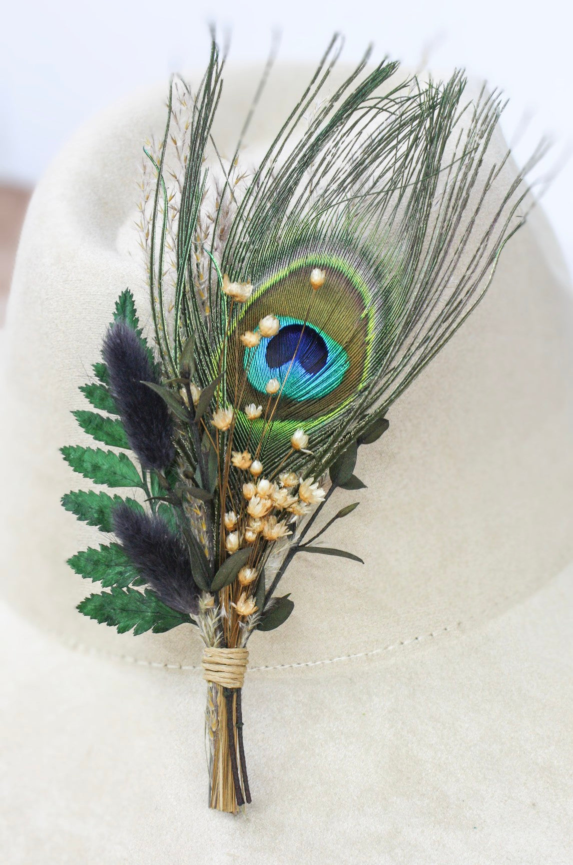 A dried floral hat pin featuring a prominent peacock feather, green leaves, purple fluffy accents, and small cream flowers, wrapped in twine and displayed on a light beige hat.