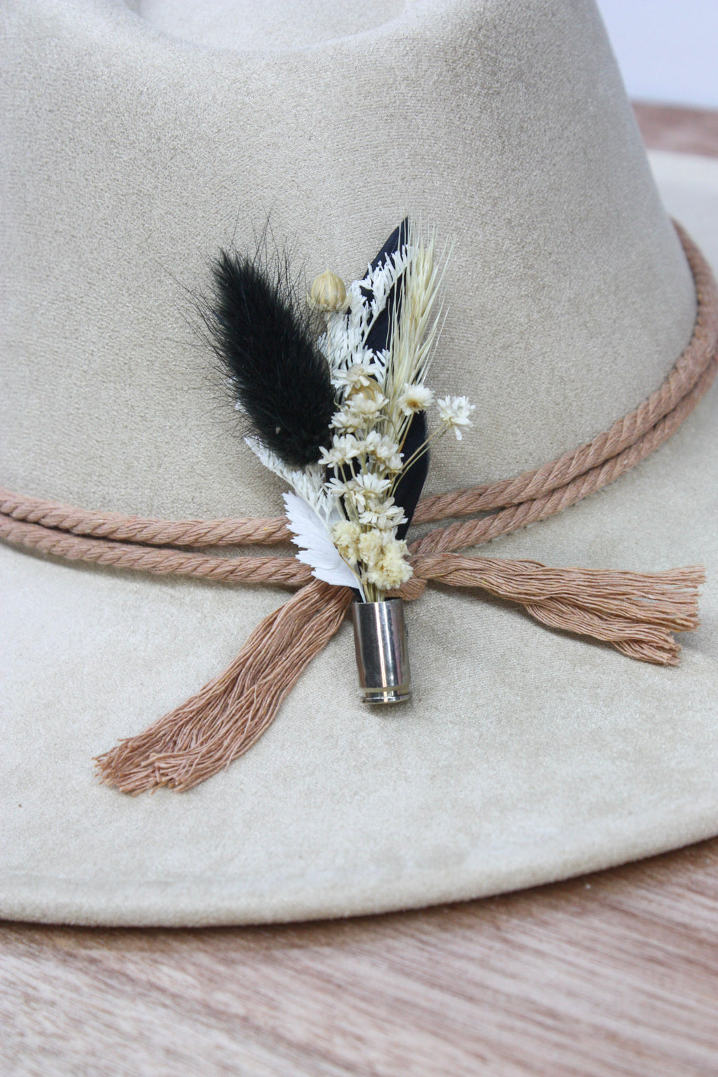 A close-up of a beige felt hat with a tan rope band and tassel, decorated with a small dried floral and feather accent in black, white, and tan, attached near the front.