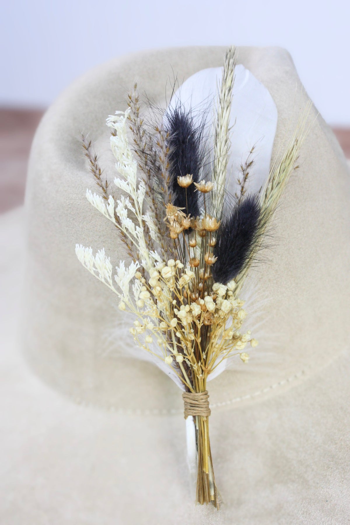 A dried floral hat pin featuring golden wheat, white baby's breath, and dark brown feather plumes, wrapped in twine and displayed on a beige felt hat.