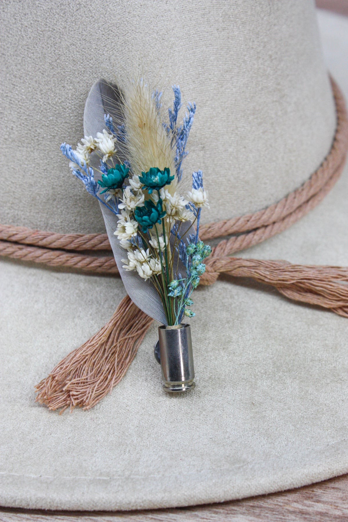 A close-up of a beige wide-brim hat with a rope band and tassel, decorated with a small floral pin featuring white and blue dried flowers, a feather, and a metallic cap.