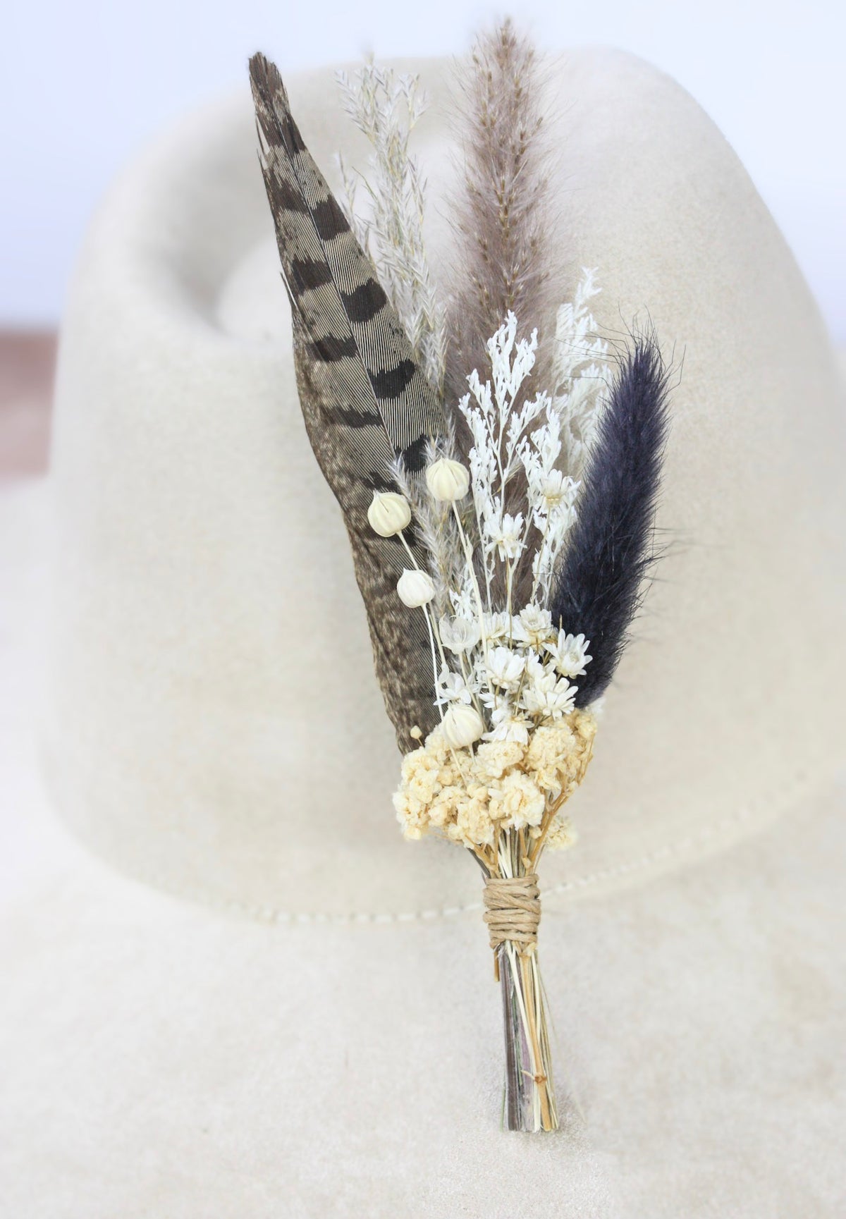 A rustic dried flower and feather hat pin displayed on a cream-colored felt hat. The bouquet features a large brown and black patterned feather, a dark navy blue feather, white baby's breath, and cream-colored flowers wrapped in natural twine.