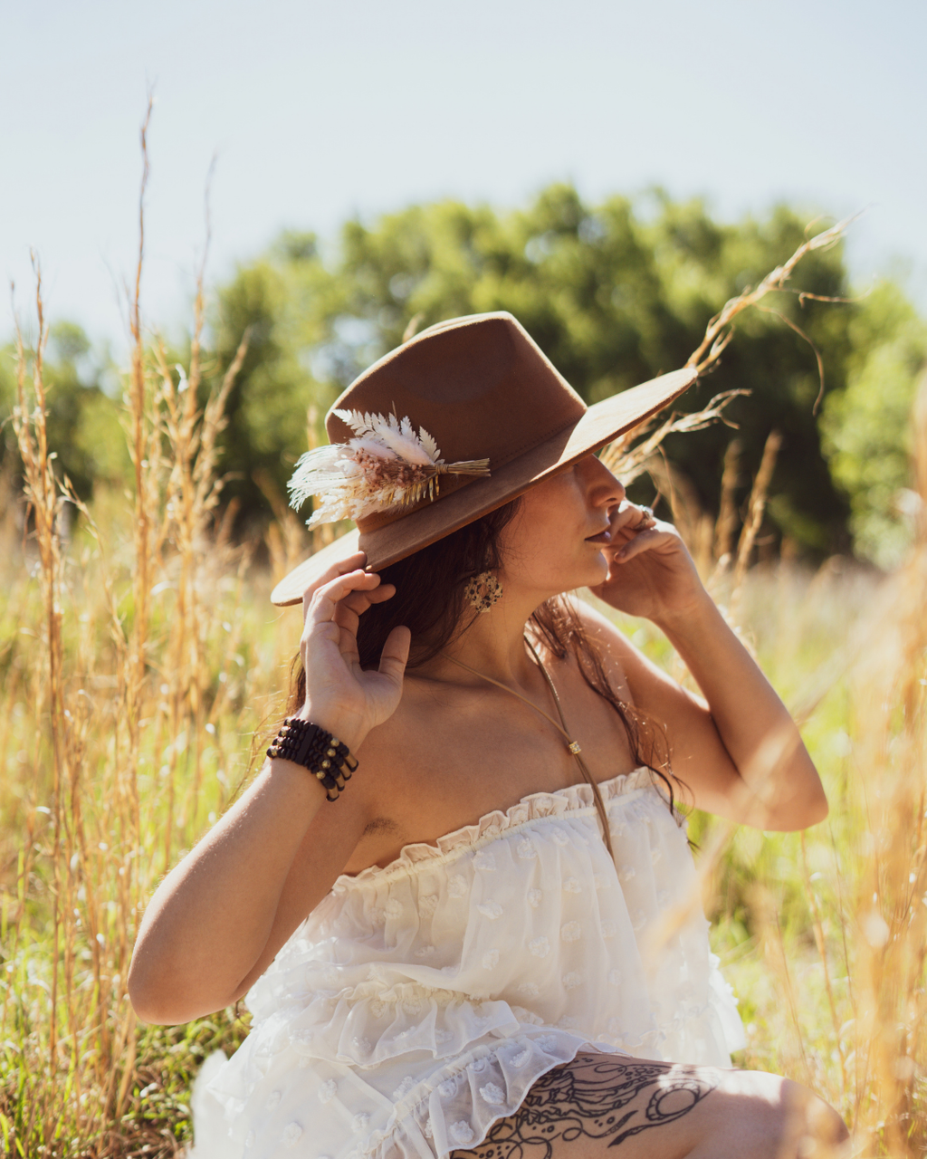A woman in a white ruffled dress sits in a sunlit grassy field, wearing a brown wide-brim hat adorned with feathers and a long necklace.