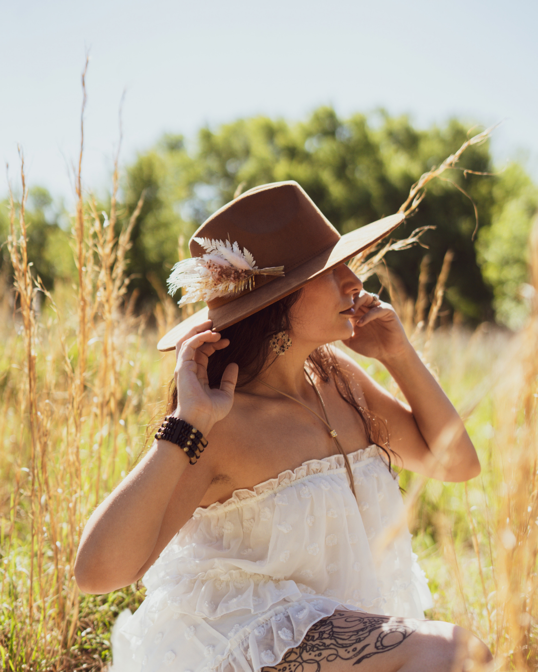 A woman in a white ruffled dress sits in a sunlit grassy field, wearing a brown wide-brim hat adorned with feathers and a long necklace.
