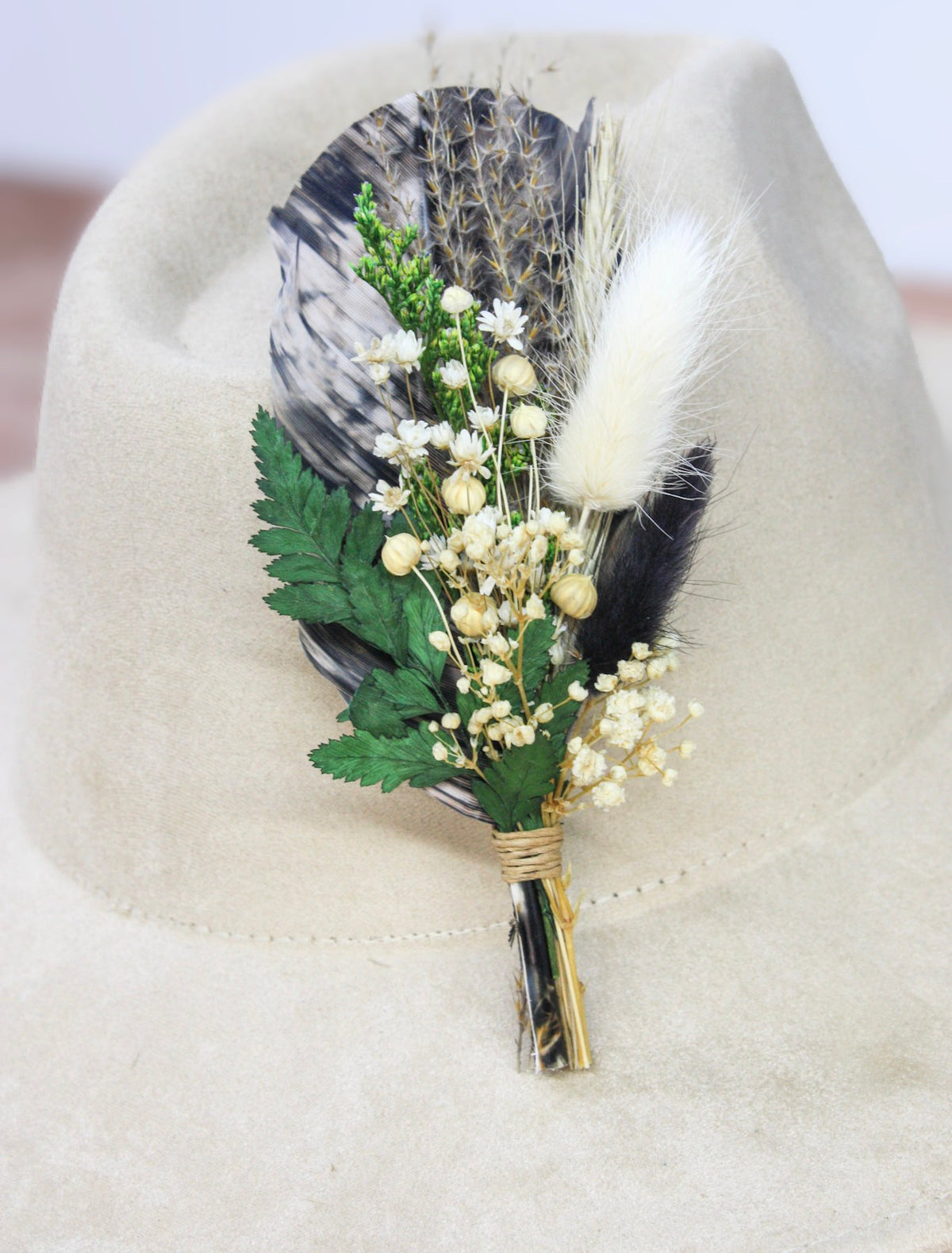 A decorative feather and floral hat pin displayed on a beige felt hat. The pin features a black and white patterned feather, a white fluffy feather, green leaves, small cream flowers, and gold-toned accents.