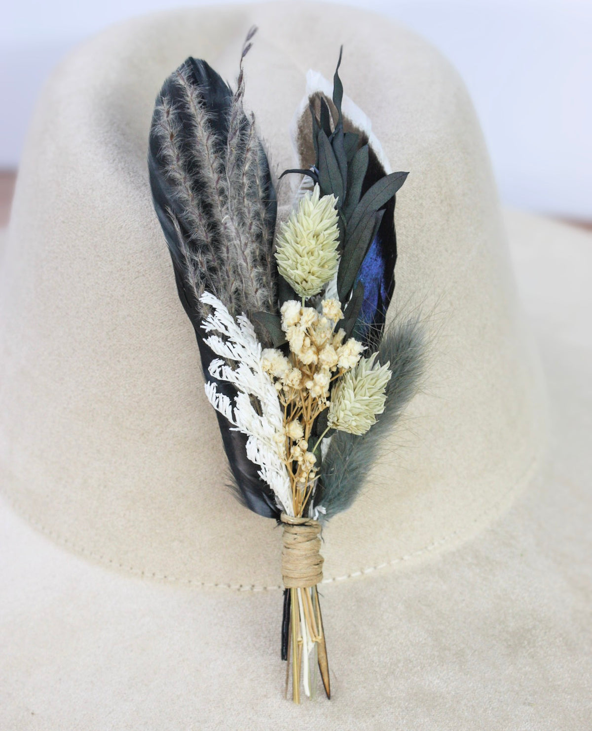 A dried floral and feather hat pin featuring black and gray feathers, cream-colored flowers, and a touch of blue, displayed on a beige fedora hat.