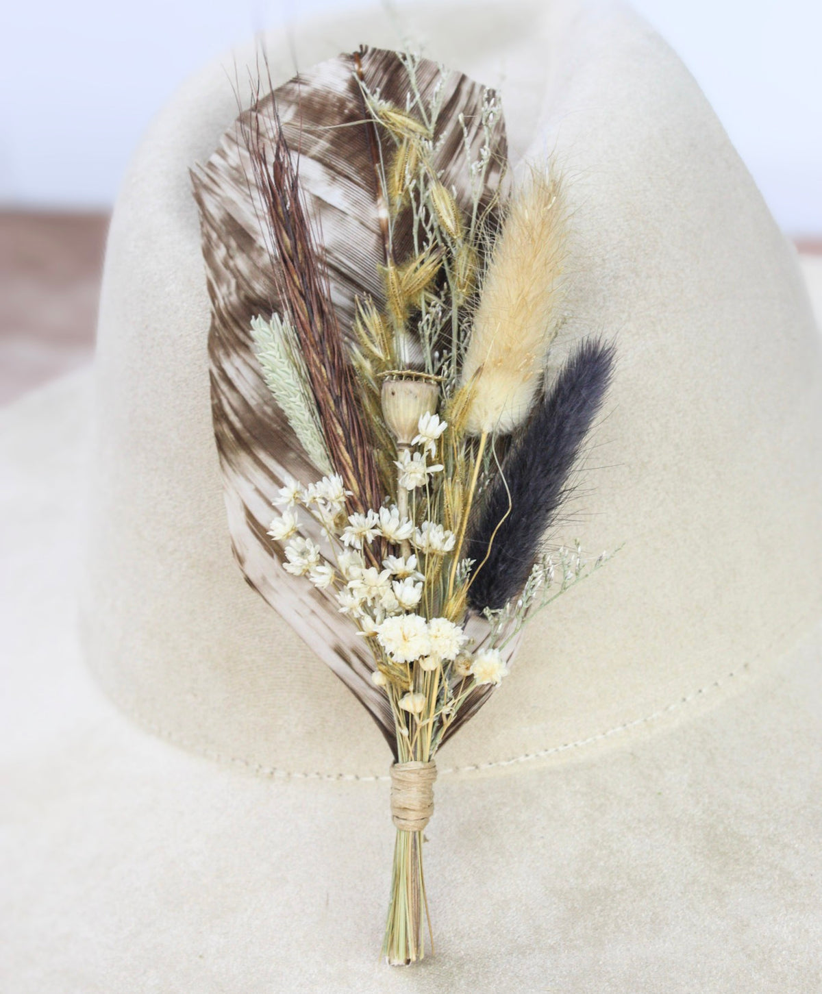 A dried floral and feather hat pin featuring brown and white speckled feathers and cream-colored flowers displayed on a beige fedora hat.