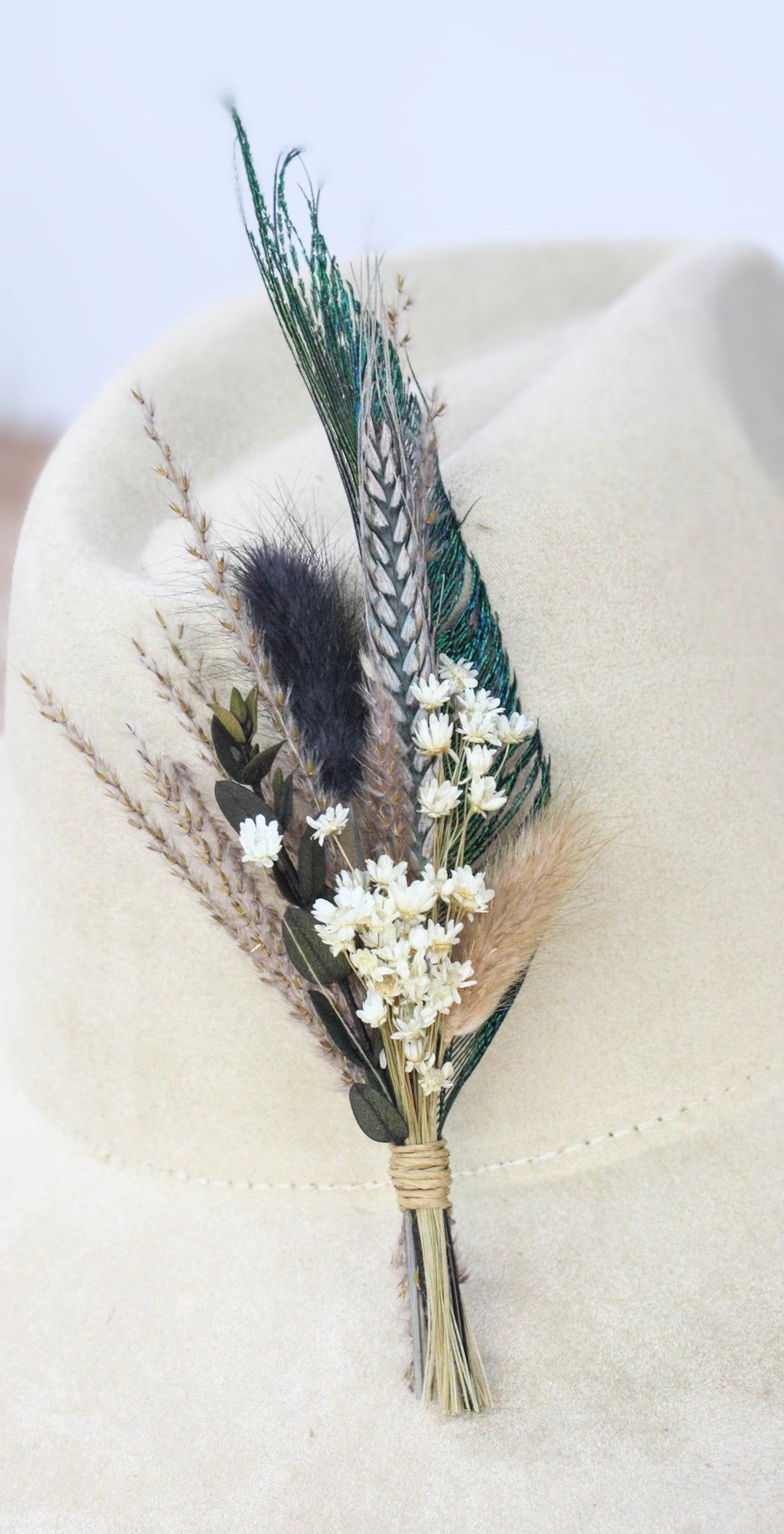 A dried floral boutonniere featuring green and brown feathers, white baby's breath, and pampas grass, displayed on a light beige felt cowboy hat.