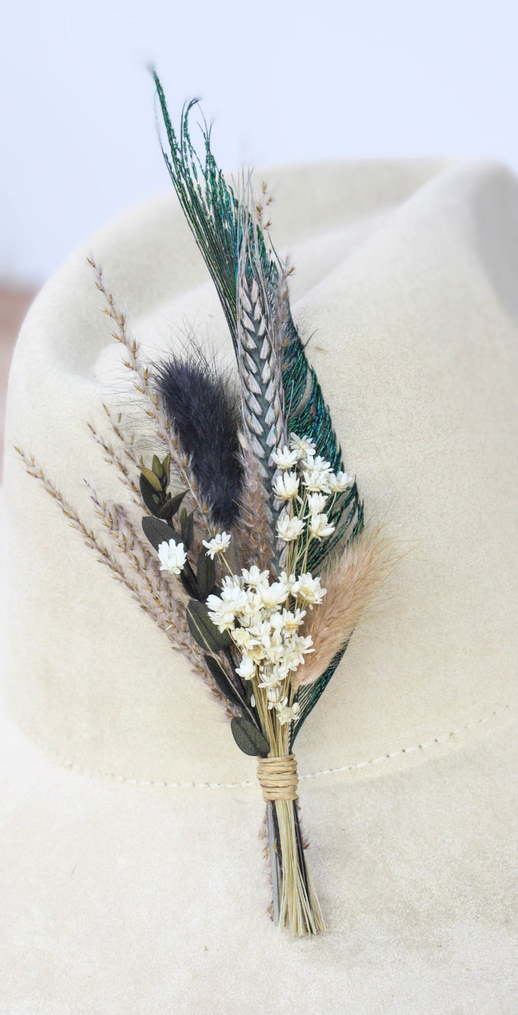 A dried floral boutonniere featuring green and brown feathers, white baby's breath, and pampas grass, displayed on a light beige felt cowboy hat.