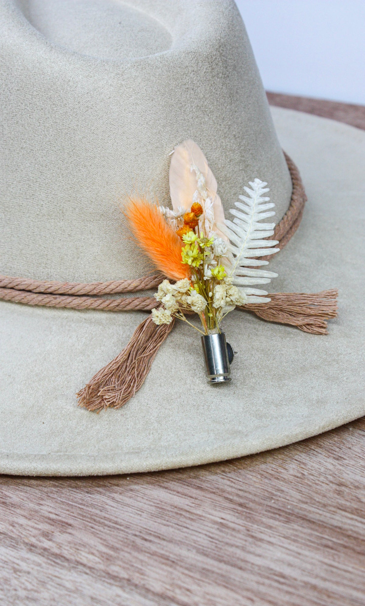 A close-up of a light beige wide-brim hat with a rope band and tassel, decorated with a small dried floral and feather accessory in orange, white, and cream tones.