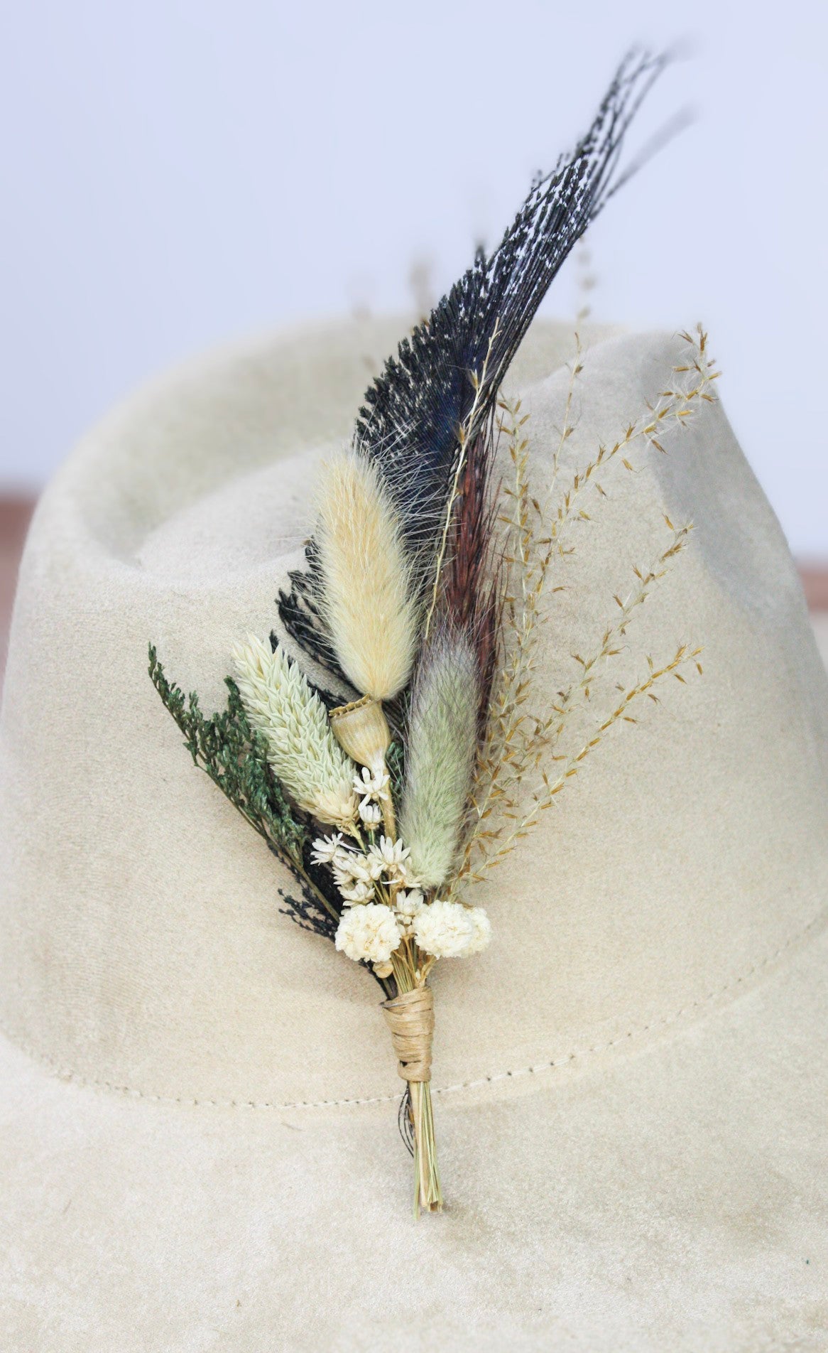 A bohemian-style dried flower and feather hat pin displayed on a light beige felt cowboy hat. The floral arrangement features cream-colored dried flowers, green and beige grasses, and dark brown and cream feathers.