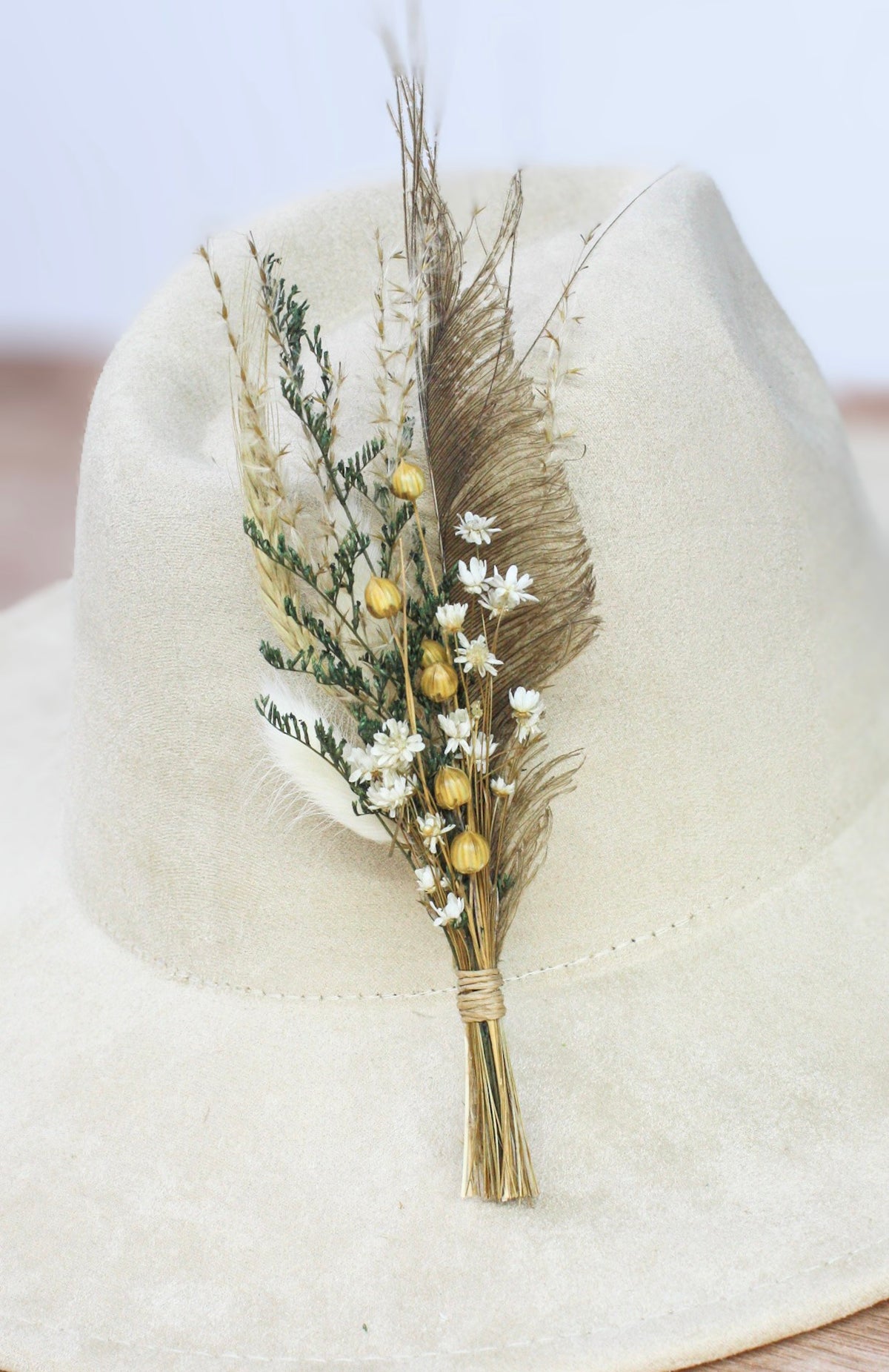 A dried floral and feather hat pin featuring cream and wheat feathers, cream-colored  and white flowers, and a touch of gold.