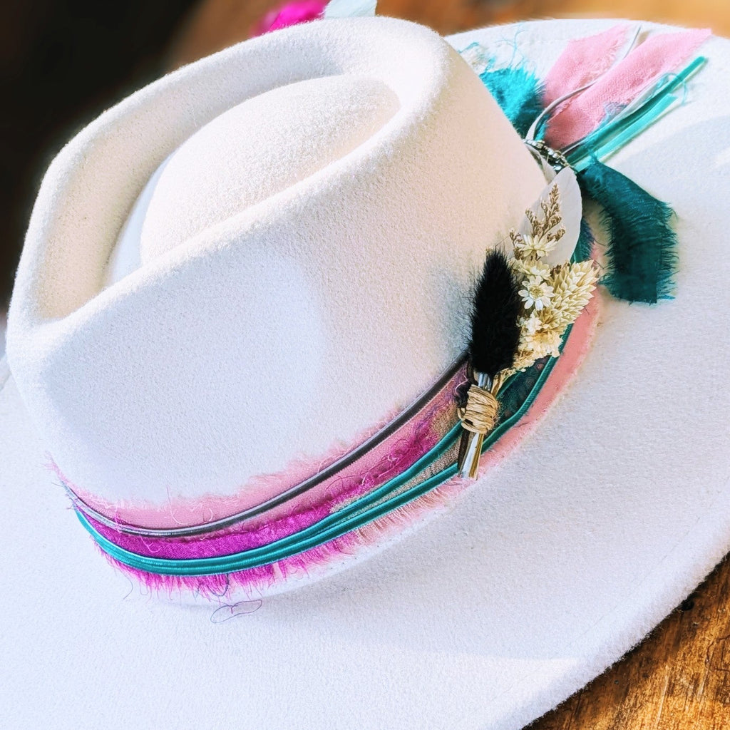 White felt hat with a pink, magenta, and teal ribbon band and a BittyBud pin of a black bunny tail, cream wheat, and white florals