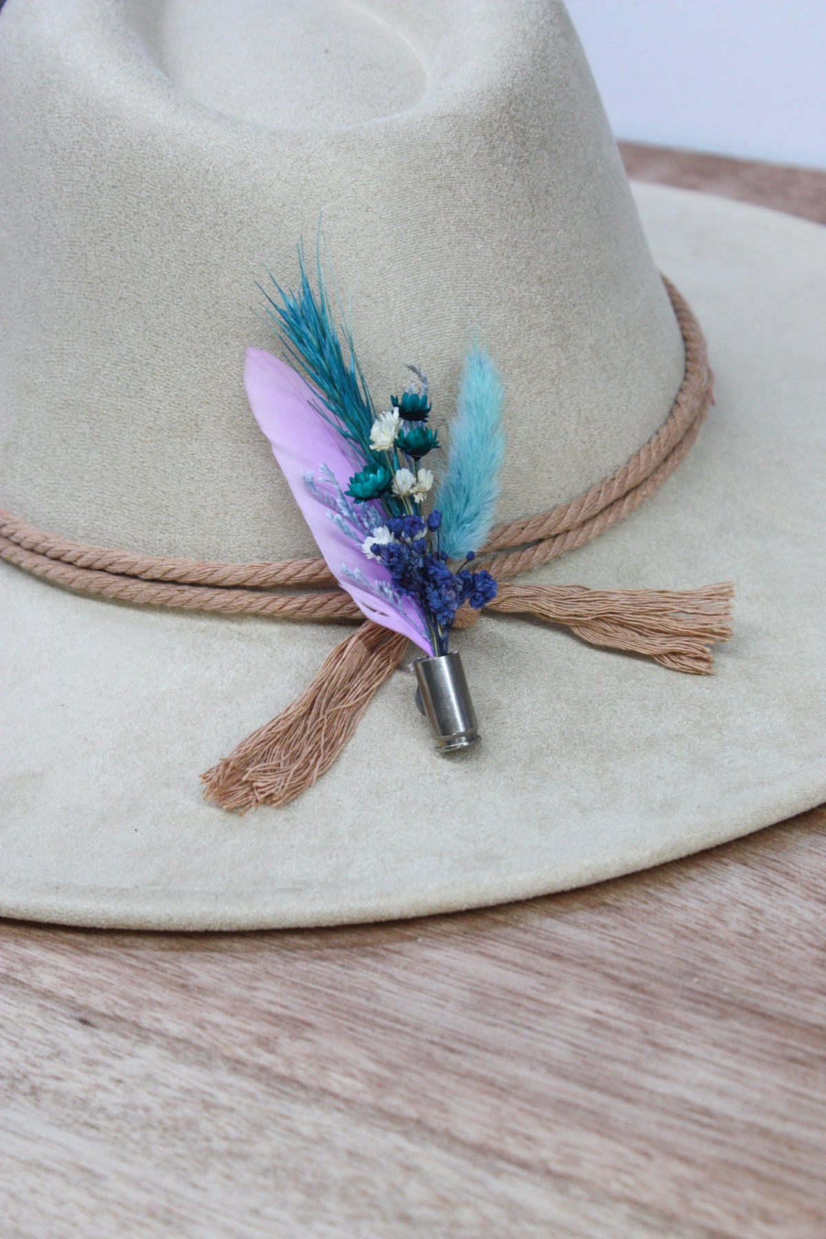 A close-up of a beige wide-brim hat with a rope band and tassels, decorated with a feathered pin featuring a lavender feather, blue feather, and small white flowers.